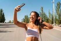 Positive female skater in rollerblades and headphones taking self shot on mobile phone blinking and sticking tongue out on sunny day in summer in city — Stock Photo