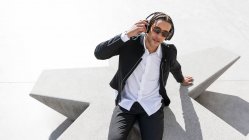 Ein junger Mann mit geflochtenem Haar, elegantem Anzug und Sonnenbrille, der Musik über drahtlose Kopfhörer genießt, während er sich auf dem sonnigen Stadtplatz ausruht — Stockfoto