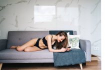 Deliziosa donna in biancheria intima nera sdraiata sul divano e messaggistica sui social media tramite telefono cellulare in accogliente salotto a casa — Foto stock