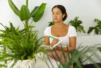 Donna etnica pensierosa con penna e agenda tra piante verdi in vaso nel giardino di casa — Foto stock