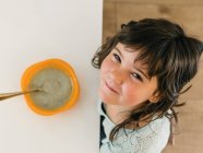 De cima de criança alegre sentada à mesa com tigela de sopa de creme e olhando para a câmera durante o almoço na cozinha em casa — Fotografia de Stock
