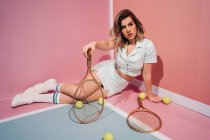 Молодая спортсменка в кроссовках и спортивной одежде сидит с теннисными ракетками и смотрит в сторону — стоковое фото