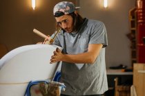 Концентрированный хипстер-мужчина с помощью инструмента писца с карандашом во время маркировки доски для сёрфинга перед формированием в мастерской — стоковое фото
