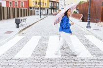Вид збоку на стильну жінку під парасолькою, що перетинає дорогу в місті і посміхається — стокове фото
