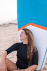 Surfista feminina alegre sentado com placa SUP azul na costa arenosa no verão e olhando para longe — Fotografia de Stock