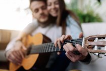 Fröhlich tätowierte männliche Musiker spielen Gitarre in der Nähe zufriedener weiblicher Geliebter, während sie einander im Sessel im Hauszimmer anschauen — Stockfoto