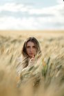 Jeune femme aux cheveux ondulés regardant la caméra dans un champ de campagne sous un ciel nuageux sur fond flou — Photo de stock