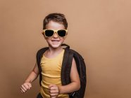 В студии классный дошкольник в солнцезащитных очках и с рюкзаком смотрит на камеру на коричневом фоне — стоковое фото