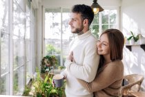 Side view of cheerful female embracing bearded male beloved with mug of coffee while looking away against window in house — Stock Photo
