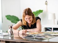 Ocupada mujer diseñadora de pintura con pincel sobre papel mientras está sentada en la mesa en la oficina creativa y trabajando en el proyecto - foto de stock