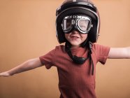 Enfant joyeux dans un casque et des lunettes de protection en regardant la caméra avec les bras tendus sur fond beige — Photo de stock