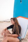Surfeuse assise avec SUP board bleu sur le bord de mer sablonneux en été et regardant loin — Photo de stock