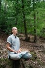 Bald man in traditional clothes sitting on rock in Lotus pose and meditating during kung fu training in forest — Stock Photo