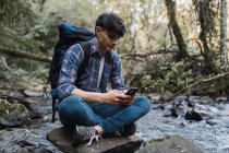 Вид сбоку мужчины-туриста с рюкзаком, сидящего у озера в одежде и просматривающего мобильный телефон во время путешествия — стоковое фото