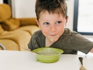 Мальчик с грязным ртом сидит за столом с миской сливочного супа во время обеда и смотрит в сторону — стоковое фото