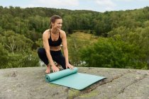 Pieno corpo donna a piedi nudi in activewear nero si sta svolgendo stuoia sulla roccia all'inizio della sessione di yoga vicino palude in natura — Foto stock