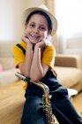 Cheerful kid in hat with saxophone looking at camera while sitting on table in house room — Stock Photo