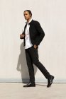 Vista lateral de cuerpo completo de contenido seguro joven étnico masculino con trenzas que usan ropa de moda y gafas de sol caminando mirando hacia la calle urbana - foto de stock
