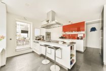 Isla de la cocina con mostrador y taburetes de bar bajo capó en moderno apartamento de espacio abierto con paredes blancas con muebles y utensilios - foto de stock