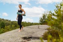 Slide view emotionless young female in black sportswear carrying rolled up mat and looking away before yoga session in lush countryside — Stock Photo