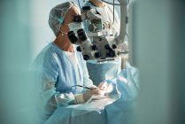 Сосредоточенная взрослая женщина-врач в стерильной маске и декоративной медицинской шапке, смотрящая через хирургический микроскоп на коллегу по урожаю в больнице — стоковое фото