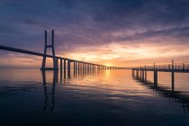 Silhouette del ponte Vasco da Gama e lungo molo situato sul tranquillo fiume Tago contro il cielo nuvoloso al tramonto in serata a Lisbona, Portogallo — Foto stock