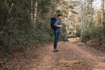 Вид сбоку путешествующего человека с рюкзаком, стоящего на песчаной дороге в лесу во время похода и отводящего взгляд — стоковое фото