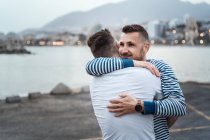 Aufrichtiger Mann umarmt bis zur Unkenntlichkeit homosexuellen Partner, während er gegen See und Berg in der Stadt wegschaut — Stockfoto