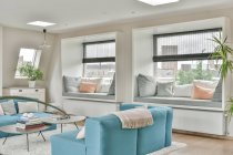 Diseño interior de sala de estar de espacio abierto con sofá azul y sillas colocadas cerca de una pequeña mesa en una alfombra suave en un apartamento moderno con paredes blancas y techo iluminado con lámparas - foto de stock