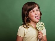 Divertente preteen bambino leccare dolce vortice lecca-lecca su sfondo verde in studio e guardando la fotocamera — Foto stock