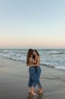 Молоді подружки обіймаються і цілуються один з одним, стоячи на піщаному пляжі біля махаючого моря на заході сонця — стокове фото