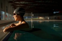 Молода красива жінка на бордюрі критого басейну, одягнена в чорний купальник, плаває у воді — стокове фото