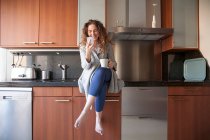 Mulher de negócios com cabelo encaracolado sentado na cozinha tomando uma infusão ao usar seu smartphone e trabalhando em casa — Fotografia de Stock