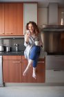 Ділова жінка з кучерявим волоссям сидить на кухні, приймаючи настій, використовуючи свій смартфон і працюючи вдома — стокове фото