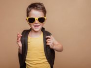 Positivo fresco preteen scolaro in occhiali da sole e con zaino guardando la fotocamera su sfondo marrone in studio — Foto stock