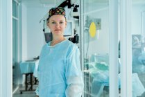 Самоуверенная взрослая женщина-врач в орнаментальной медицинской шапке смотрит на камеру напротив стеклянной стены в больнице — стоковое фото