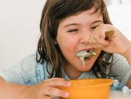Adorable enfant mangeant une délicieuse soupe à la crème dans un bol en plastique assis à table pendant le déjeuner à la maison — Photo de stock