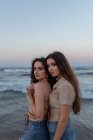 Young girlfriends embracing each other while standing on sandy beach near waving sea at sundown ooking at camera — Stock Photo