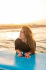 Surfista feminina deitada na prancha SUP e flutuando na água calma do mar no dia ensolarado olhando para longe durante o pôr do sol — Fotografia de Stock
