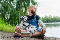 Pensive bald male with beard in casual outfit sitting with obedient spotted Great Dane puppy on lake with green trees on summer day — Stock Photo