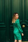 Весела жінка в стильній зеленій сукні, що стоїть біля декоративних дерев'яних дверей на вулиці і дивиться на камеру — стокове фото
