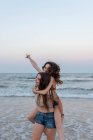 Mulher nova que dá o passeio do piggyback à namorada quando estando na água do mar acenando na noite — Fotografia de Stock