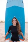 Mujer surfista feliz de pie con tabla SUP azul en la orilla del mar de arena en verano y mirando a la cámara - foto de stock