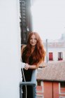 Content female with red hair standing on balcony with cup of beverage and looking at camera — Stock Photo