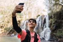 Lächelnder männlicher Wanderer, der beim Trekking auf dem Hintergrund von Wasserfall und See im Wald Selbstaufnahmen mit dem Smartphone macht — Stockfoto