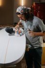 Male shaper using electric planer and polishing surface of surfboard in workshop — Stock Photo