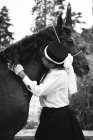 Hembra negra adulta seria en sombrero y elegante atuendo con guantes acariciando caballos marrones cerca de plantas verdes y árboles en el campo - foto de stock