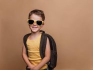 Positivo legal pré-adolescente estudante em óculos de sol e com mochila olhando para a câmera no fundo marrom em estúdio — Fotografia de Stock