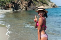 Вид збоку весела етнічна жінка-туристка в купальнику і капелюсі, дивлячись на камеру на узбережжі океану, поки вона робить символ перемоги — стокове фото