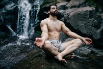 Friedliches Männchen ohne Hemd sitzt in Padmasana mit Mudra-Händen und geschlossenen Augen, während es Yoga macht und auf nassen Felsen in der Nähe des Wasserfalls meditiert — Stockfoto